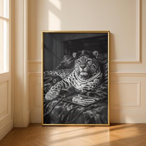 May include: Black and white framed print of a leopard wearing a sleep mask and pearl necklace, reclining on a bed. A tray with pearls sits in front of the leopard. The print has a gold frame and is displayed in a room with a wooden floor.