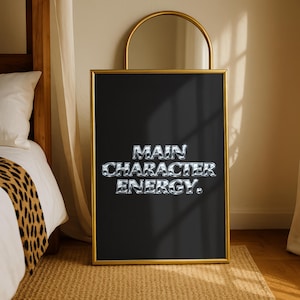Op de afbeelding: Een zwart ingelijste poster met de woorden "MAIN CHARACTER ENERGY" in zilveren, metallic letters. De lijst is goudkleurig en de poster wordt tentoongesteld op een geweven vloerkleed. Boven de poster bevindt zich een goudkleurige spiegel.