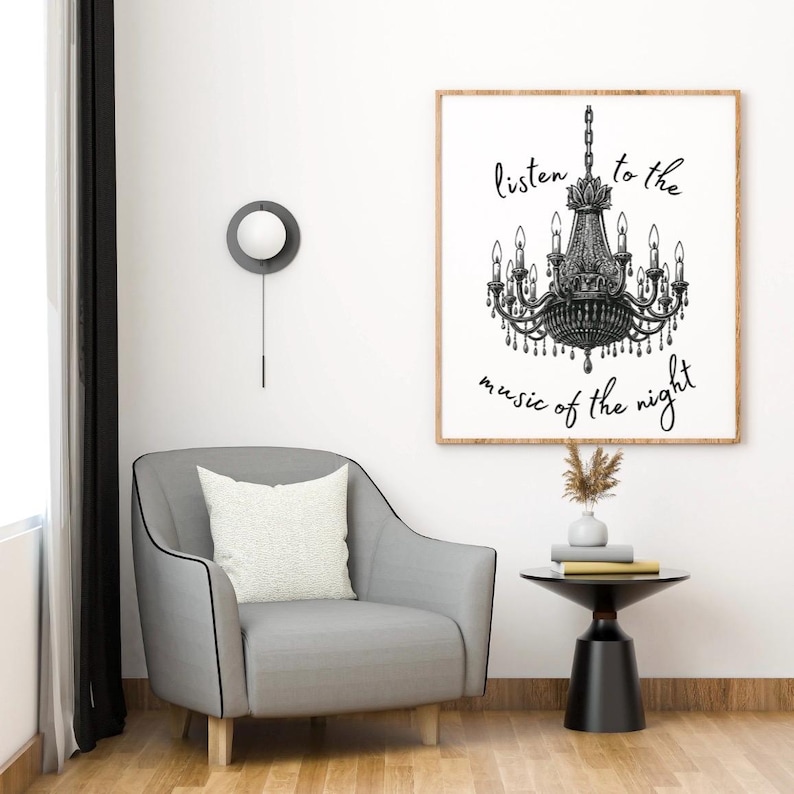 May include: A framed print with a black and white chandelier illustration and the text "listen to the music of the night." The print hangs above a gray armchair with a white pillow. A small black side table holds books and a vase.