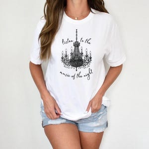 May include: White t-shirt featuring a black chandelier graphic with the text "listen to the music of the night". The shirt has short sleeves and a crew neck.