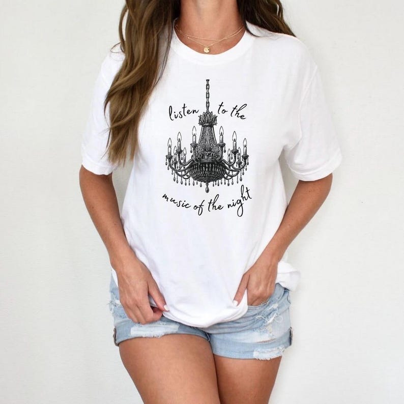 May include: White t-shirt featuring a black chandelier graphic with the text "listen to the music of the night". The shirt has short sleeves and a crew neck.