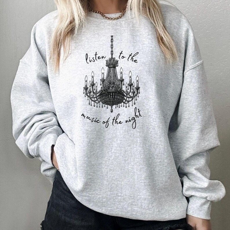 May include: A light gray sweatshirt featuring a black and white chandelier design. The text "listen to the music of the night" is printed above and below the chandelier. The sweatshirt has long sleeves and a crew neck.