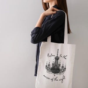 May include: Off-white tote bag featuring a black chandelier illustration and the text "listen to the music of the night." The bag has long straps and is carried over the shoulder. The bag is made of a light-colored fabric.