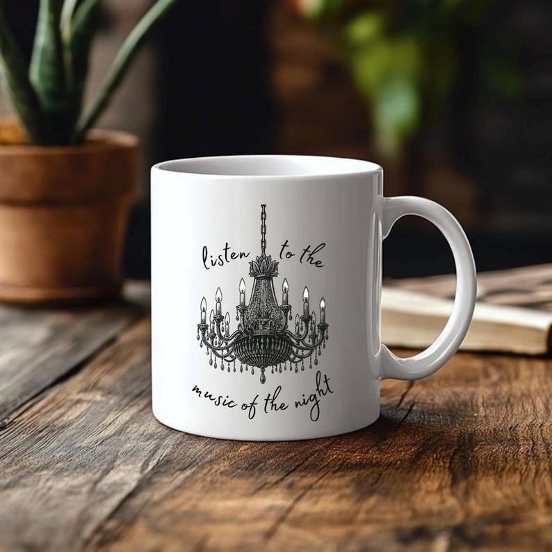 May include: White ceramic mug featuring a black chandelier design with the text "listen to the music of the night". The mug has a curved handle and is set on a wooden surface.