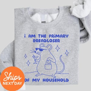 May include: A light gray crewneck sweatshirt featuring a blue graphic of a mouse wearing sunglasses, holding a martini glass and a small bag. The text reads: I AM THE PRIMARY BREADLOSER OF MY HOUSEHOLD, surrounded by small four-pointed star icons.