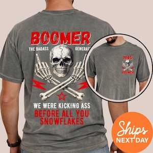 May include: Gray t-shirt with a skull graphic, skeleton hands, and the word "BOOMER" in red. The shirt also features the text "WE WERE KICKING ASS BEFORE ALL YOU SNOWFLAKES". A smaller graphic is on the upper left chest.