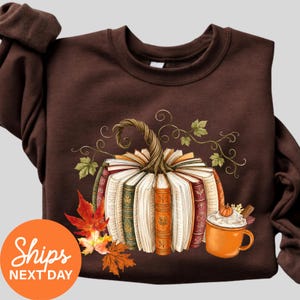 May include: A dark brown long-sleeve sweatshirt featuring a graphic of a pumpkin constructed from open books with colorful spines. The design includes green vines, orange and red autumn leaves, and an orange mug filled with a creamy beverage.