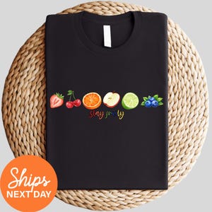 May include: A black t-shirt featuring a horizontal row of colorful fruit illustrations including a strawberry, cherries, an orange slice, an apple slice, a lime slice, and blueberries, with the text "slay fruity" written in a rainbow-colored script.