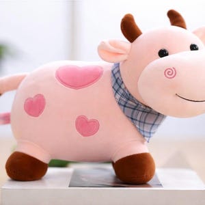 Puede incluir: Un peluche de vaca rosa con pezuñas, cuernos y puntas de orejas marrones. La vaca tiene manchas rosas en forma de corazón y una bufanda a cuadros azul. El juguete tiene una cara sonriente.
