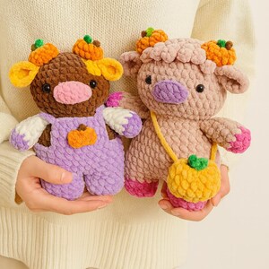May include: Two handmade crochet cow plushies. One is brown and purple, wearing overalls with a pumpkin applique. The other is pink, with a yellow purse. Both have pumpkin crowns and are held in front of a cream sweater.