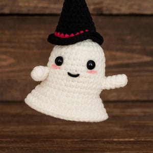 May include: A handmade, white crochet ghost wearing a black witch hat with a red trim. The ghost has black button eyes, rosy cheeks, and a smiling mouth. This Halloween decoration is suspended against a wooden background.