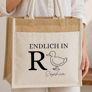 K&ouml;nnte beinhalten: Eine quadratische Jute-Einkaufstasche mit zwei Tragegriffen und einer cremefarbenen Canvas-Vordertasche. Die Tasche zeigt den schwarzen Aufdruck ENDLICH IN, einen gro&szlig;en Buchstaben R, eine Strichzeichnung einer Ente und den Namen Sophia in Schreibschrift.