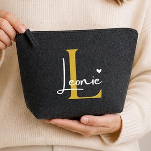 May include: A dark grey felt zipped pouch featuring a large gold letter L and the name Leonie printed in white script with a small white heart icon. The pouch has a trapezoidal shape and a matching dark grey zip pull tab on the top right.