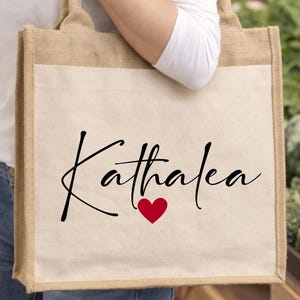 Peut inclure: Un sac fourre-tout beige et blanc avec le nom "Kathalea" en écriture noire et un cœur rouge. Le sac est fait d'un matériau semblable à du jute avec un panneau avant blanc. Le sac est porté par une personne.