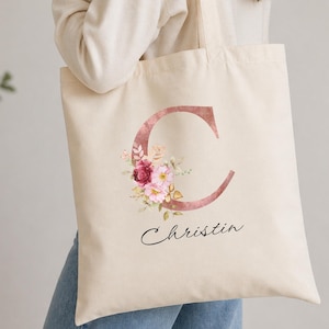 Puede incluir: Bolso tote color crema con una gran letra "C" en oro rosa y un diseño floral de flores rosas y rojas. El nombre "Christin" está escrito en cursiva negra debajo. El bolso tiene asas largas.