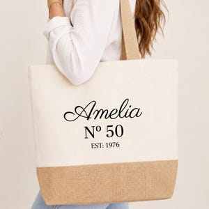 Könnte beinhalten: Eine Canvas-Tragetasche mit einem naturfarbenen beigen Boden und Trägern. Der Hauptteil ist cremefarben mit dem Namen "Amelia" in Schreibschrift, zusammen mit "Nº 50" und "EST: 1976" in Schwarz gedruckt.