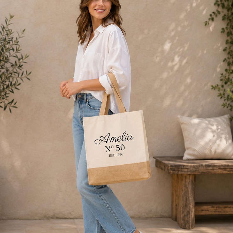 May include: A beige tote bag with a natural jute bottom and handles. The bag features the name "Amelia" in a script font, with "No 50" and "EST. 1976" printed below in black.