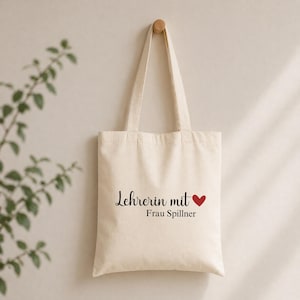 May include: Cream-colored tote bag with black script text that reads "Lehrerin mit" and a red heart, followed by "Frau Spillner". The bag hangs from a wooden peg on a light-colored wall. The bag has two handles.