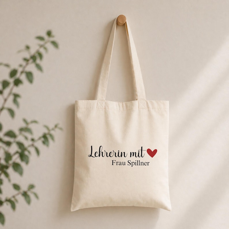 May include: Cream-colored tote bag with black script text that reads "Lehrerin mit" and a red heart, followed by "Frau Spillner". The bag hangs from a wooden peg on a light-colored wall. The bag has two handles.