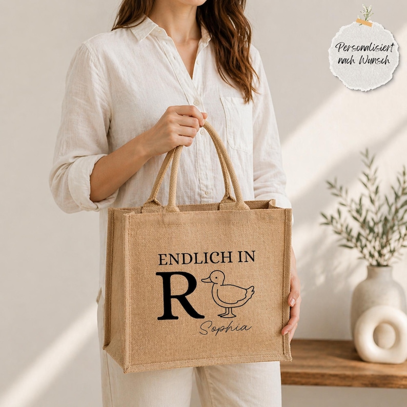 Könnte beinhalten: Eine rechteckige Tragetasche aus beigem Jute-Stoff mit zwei Griffen. Auf der Tasche steht der Text "ENDLICH IN R", eine Entenillustration und der Name "Sophia". Die Tasche wird von einer Person gehalten.