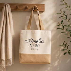 May include: A beige tote bag with a natural jute bottom and handles. The bag features the name "Amelia" in elegant script, with "N&ordm; 50" and "EST: 1976" printed below. The bag is hanging on a wooden rack.