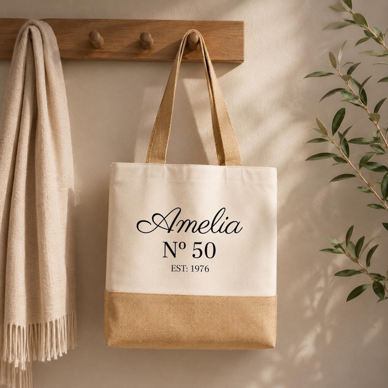 May include: A beige tote bag with a natural jute bottom and handles. The bag features the name "Amelia" in elegant script, with "N&ordm; 50" and "EST: 1976" printed below. The bag is hanging on a wooden rack.
