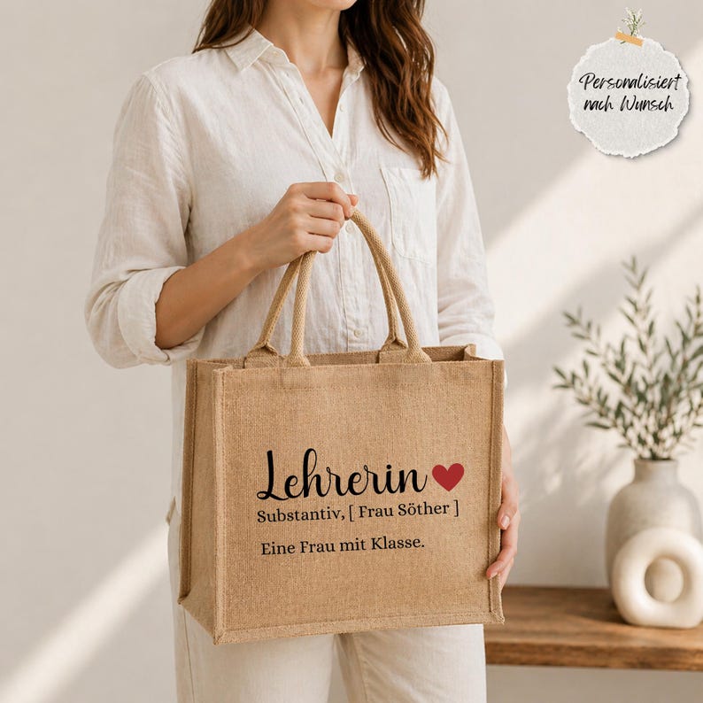 May include: A rectangular tan jute tote bag with two handles. The front features black printed text that reads "Lehrerin" followed by a red heart, "Substantiv, [ Frau S&ouml;ther ]", and "Eine Frau mit Klasse." A small white circular tag reads "Personalisiert nach Wunsch."
