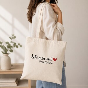 May include: A cream-colored tote bag with the text "Lehrerin mit" and "Frau Spillner" in black script, a red heart symbol, and two cream-colored handles. The bag is made of a sturdy fabric.