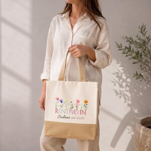 May include: A cream-colored canvas tote bag with a tan bottom panel. The front features a colorful illustration of various wildflowers above the text RENTNERIN and the script text Sabine seit 2026. The bag has two matching canvas shoulder straps.