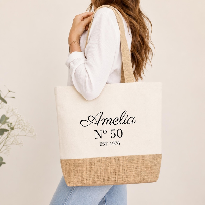May include: A canvas tote bag with a natural beige bottom and straps. The main body is off-white with the name "Amelia" in a cursive font, along with "N&ordm; 50" and "EST: 1976" printed in black.