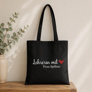 May include: Black tote bag with the text "Lehrerin mit Frau Spillner" in white script, a red heart, and black handles. The bag is made of a sturdy material and is designed for carrying items.
