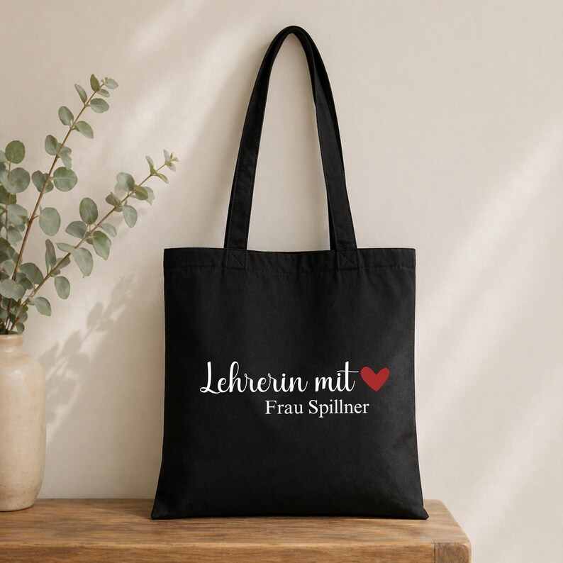 May include: Black tote bag with the text "Lehrerin mit Frau Spillner" in white script, a red heart, and black handles. The bag is made of a sturdy material and is designed for carrying items.