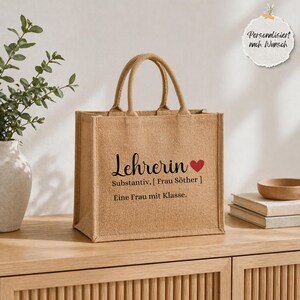 May include: A tan jute tote bag with two handles, featuring black printed text that reads "Lehrerin," a red heart icon, and the German text "Substantiv, [ Frau S&ouml;ther ] Eine Frau mit Klasse." The bag has a structured, rectangular shape.