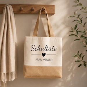 May include: A beige tote bag with a natural burlap bottom and handles. The bag features the text "Schultüte" above a heart and a line, with "FRAU MÜLLER" below. The bag is hanging on a wooden rack with a beige scarf to the left.