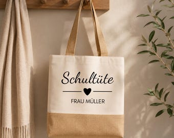 May include: A beige tote bag with a natural burlap bottom and handles. The bag features the text "Schultüte" above a heart and a line, with "FRAU MÜLLER" below. The bag is hanging on a wooden rack with a beige scarf to the left.