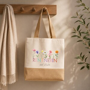 May include: A beige canvas tote bag with a brown burlap base. The front features a colorful floral illustration above the text RENTNERIN and the year 2026. The bag has two long shoulder straps and a rectangular shape with a clean, simple design.