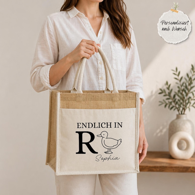 K&ouml;nnte beinhalten: Eine quadratische Jute-Einkaufstasche mit zwei Tragegriffen und einer cremefarbenen Canvas-Vordertasche. Die Tasche zeigt den schwarzen Aufdruck ENDLICH IN, einen gro&szlig;en Buchstaben R, eine Strichzeichnung einer Ente und den Namen Sophia in Schreibschrift.