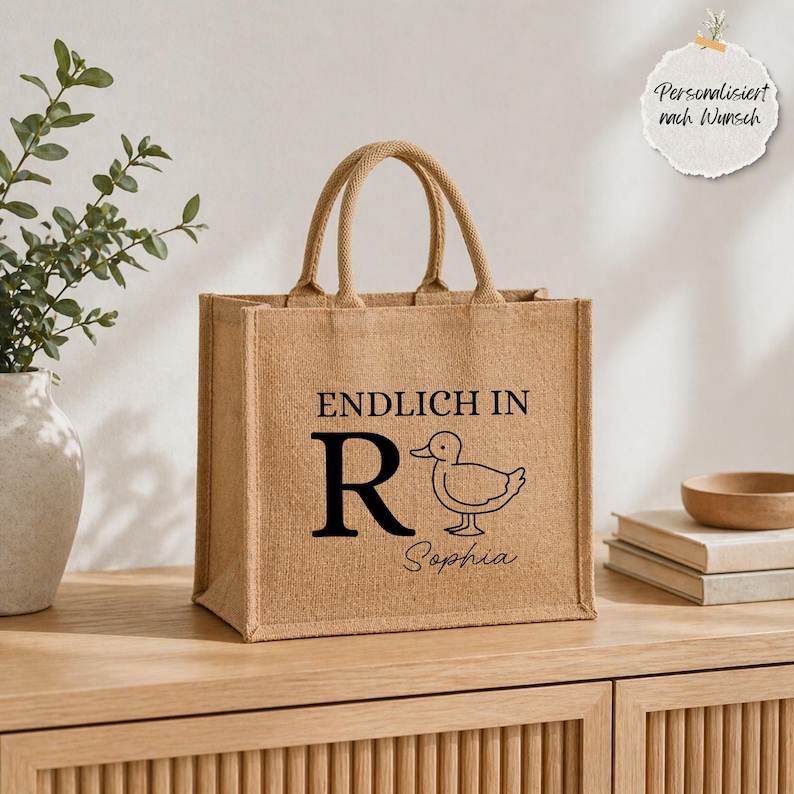 K&ouml;nnte beinhalten: Eine beigefarbene Jute-Tragetasche mit zwei Henkeln. Auf der Vorderseite befindet sich der schwarze Schriftzug ENDLICH IN, ein gro&szlig;er Buchstabe R, eine Strichzeichnung einer Ente und der Name Sophia in Schreibschrift. Ein rundes Etikett zeigt Personalisiert nach Wunsch.