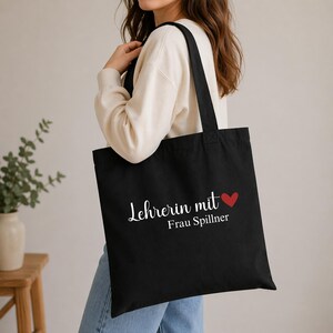 May include: Black tote bag with white text that reads "Lehrerin mit Frau Spillner" and a red heart. The bag has two black handles and is carried over the shoulder.