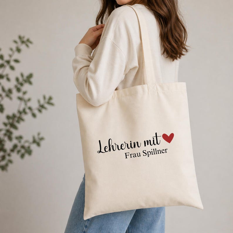 May include: Cream-colored tote bag with black script text that reads "Lehrerin mit" and a red heart, followed by "Frau Spillner". The bag has long straps and is made of a sturdy fabric, perfect for carrying books or groceries.