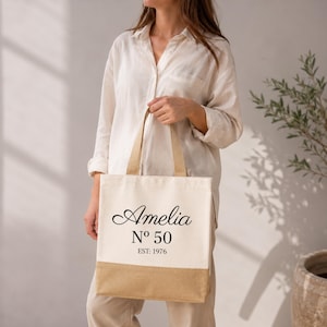 May include: Cream-colored tote bag with a natural jute bottom and handles. The bag features the name "Amelia" in elegant script, along with "N&ordm; 50" and "EST: 1976" in black text. A stylish and personalized accessory.