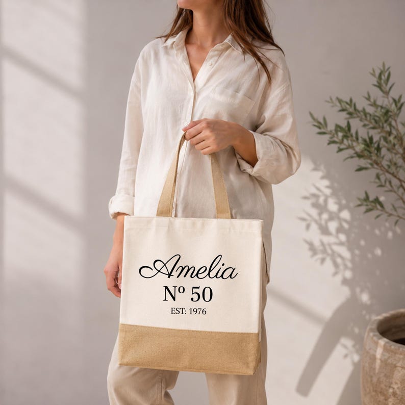 May include: Cream-colored tote bag with a natural jute bottom and handles. The bag features the name "Amelia" in elegant script, along with "N&ordm; 50" and "EST: 1976" in black text. A stylish and personalized accessory.