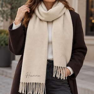 May include: A cream-colored scarf with a subtle herringbone weave pattern and long fringe at both ends. The name "Hanna" is embroidered in a black cursive script on the lower portion of the scarf, which is worn over a white top and dark coat.