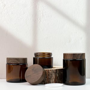 Könnte beinhalten: Drei bernsteinfarbene Glasbehälter mit Holzdeckeln. Ein Behälter hat seinen Deckel abgenommen, wodurch die Öffnung sichtbar wird. Die Behälter sind unterschiedlich groß und stehen auf einer weißen Oberfläche. Ein Behälter steht auf einer kleinen Holzplattform.