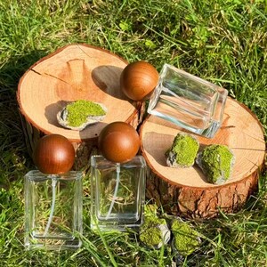 Puede incluir: Dos frascos de perfume de vidrio transparente con tapones esféricos de madera se exhiben sobre una rodaja de madera. Los frascos están rodeados de pequeñas rocas cubiertas de musgo verde y colocados sobre hierba verde. La escena está iluminada por luz natural.