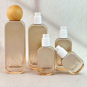 Könnte beinhalten: Eine Sammlung rechteckiger, klarer Glasflaschen mit abgerundeten Ecken. Die Flaschen haben eine goldfarbene Tönung und weiße Pumpspender. Eine Flasche hat eine Holzkugel als Verschluss. Die Flaschen sind unterschiedlich groß.