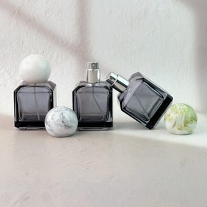 Könnte beinhalten: Zwei quadratische Parfümflakons aus dunkelgrauem Glas mit silbernen Sprühköpfen. Ein Flakon hat einen soliden weißen kugelförmigen Verschluss, während der andere auf der Seite liegt. Zwei dekorative Steinkugeln, eine weiß mit grauer Marmorierung und eine grün mit weißer Marmorierung, liegen davor.