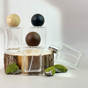 May include: Clear glass perfume bottles with wooden and black spherical caps are displayed on a rustic wooden base. The bottles are rectangular with a spray nozzle. Green moss accents the base, creating a natural aesthetic.