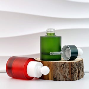 以下が含まれることがあります： 赤と緑のガラス製スポイトボトルが2つ、木の輪切りに置かれています。緑色のボトルは直立しており、スポイトが部分的に挿入されています。赤いボトルは横になっています。ボトルはマット仕上げです。