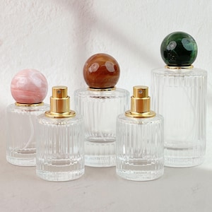 Pode incluir: Cinco frascos de perfume de vidro transparente com borrifadores dourados e tampas esféricas decorativas de pedra. As tampas são rosa, marrons e verdes. Os frascos têm um design canelado e estão dispostos sobre uma superfície branca.